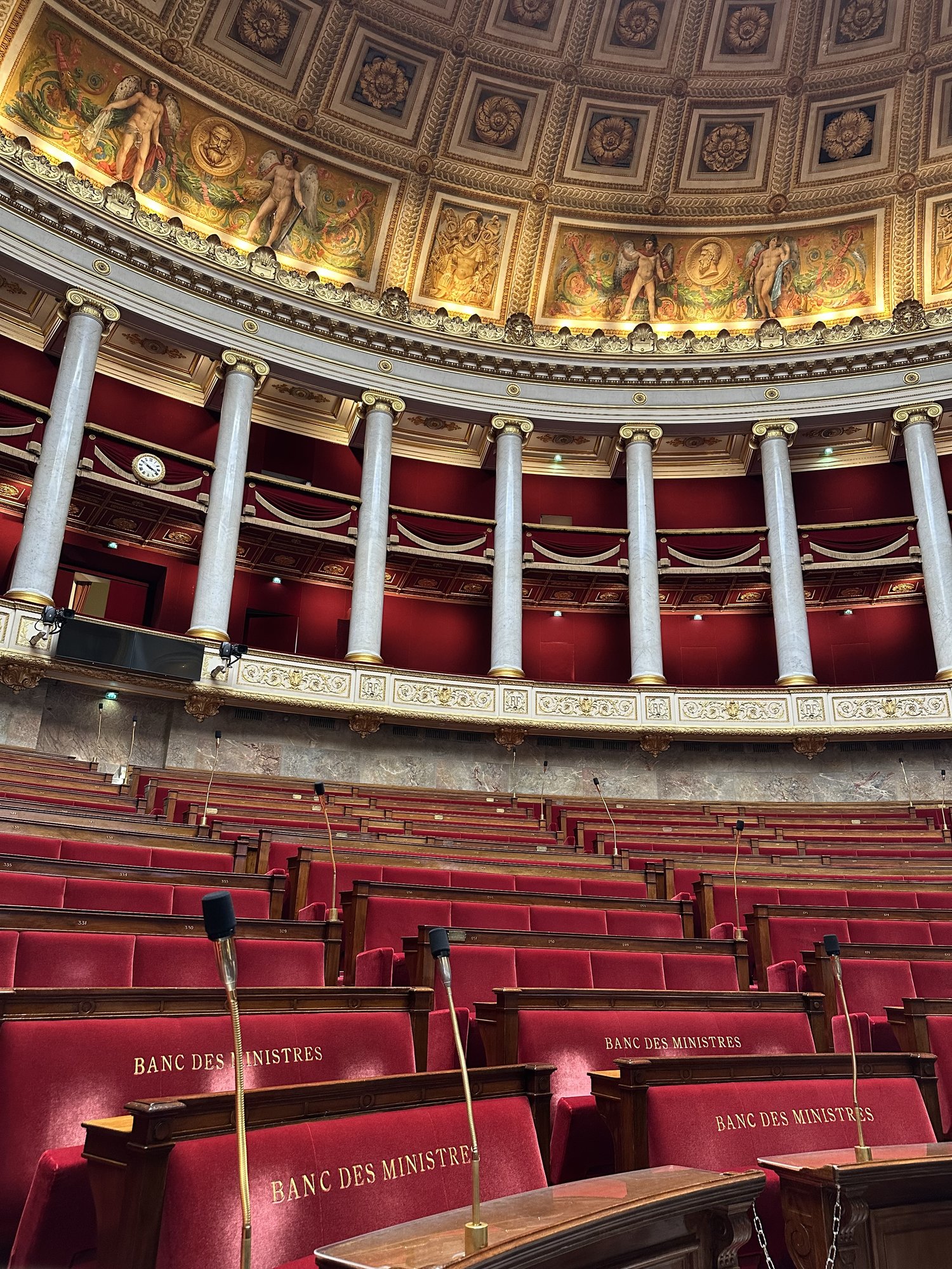 Photo de l'hémicycle de l'Assemblée nationale française en mars 2025. Les bancs vides révèlent le découpage politique en blocs : la gauche (NFP) à gauche du président, le centre au milieu, la droite et le RN à droite.