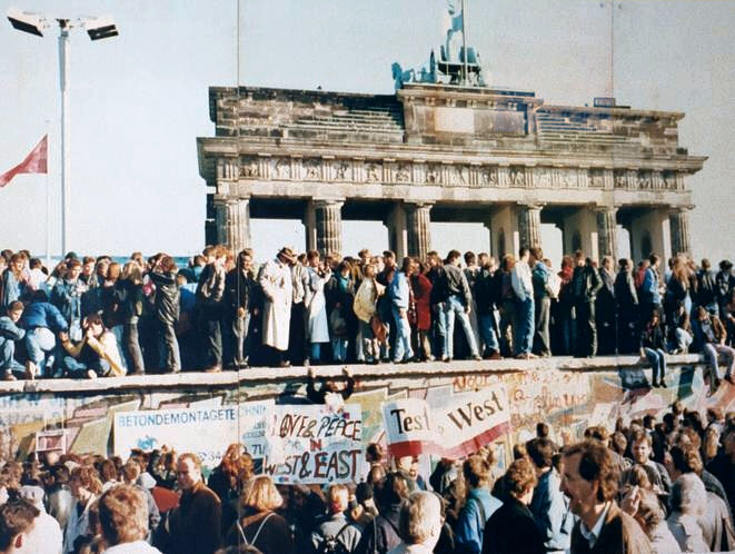 Photographie d'archive en couleurs : des dizaines d'Allemands de l'Est et de l'Ouest grimpent et se rassemblent sur le mur de Berlin devant la Porte de Brandebourg illuminée, dans la nuit de novembre 1989.