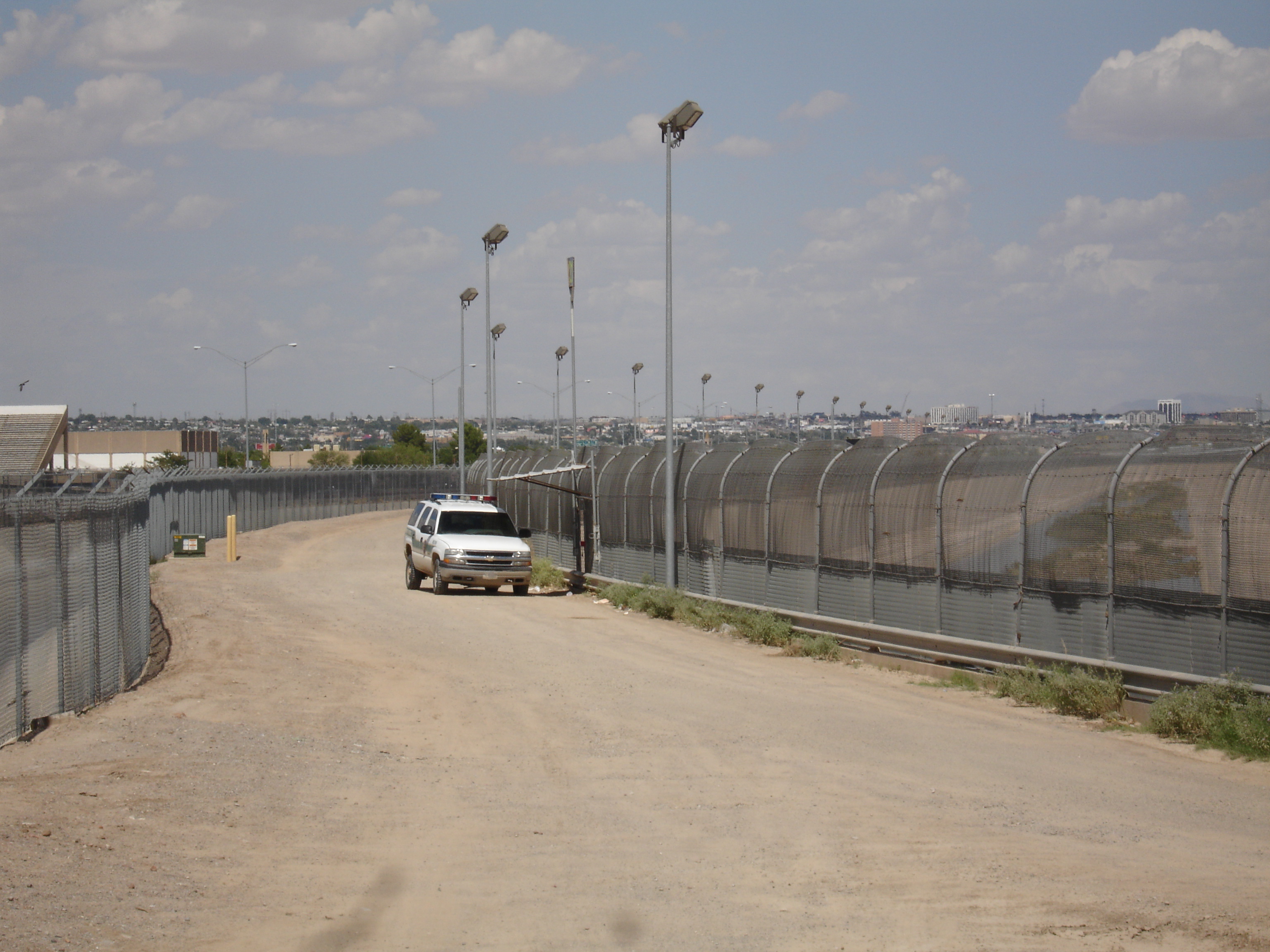Vue rapprochée d'une portion du mur frontalier entre les États-Unis et le Mexique, dans le secteur d'El Paso au Texas. Une haute clôture métallique aux barreaux serrés s'étire en ligne droite à travers une zone aride. Une route de service longe le mur du côté américain.