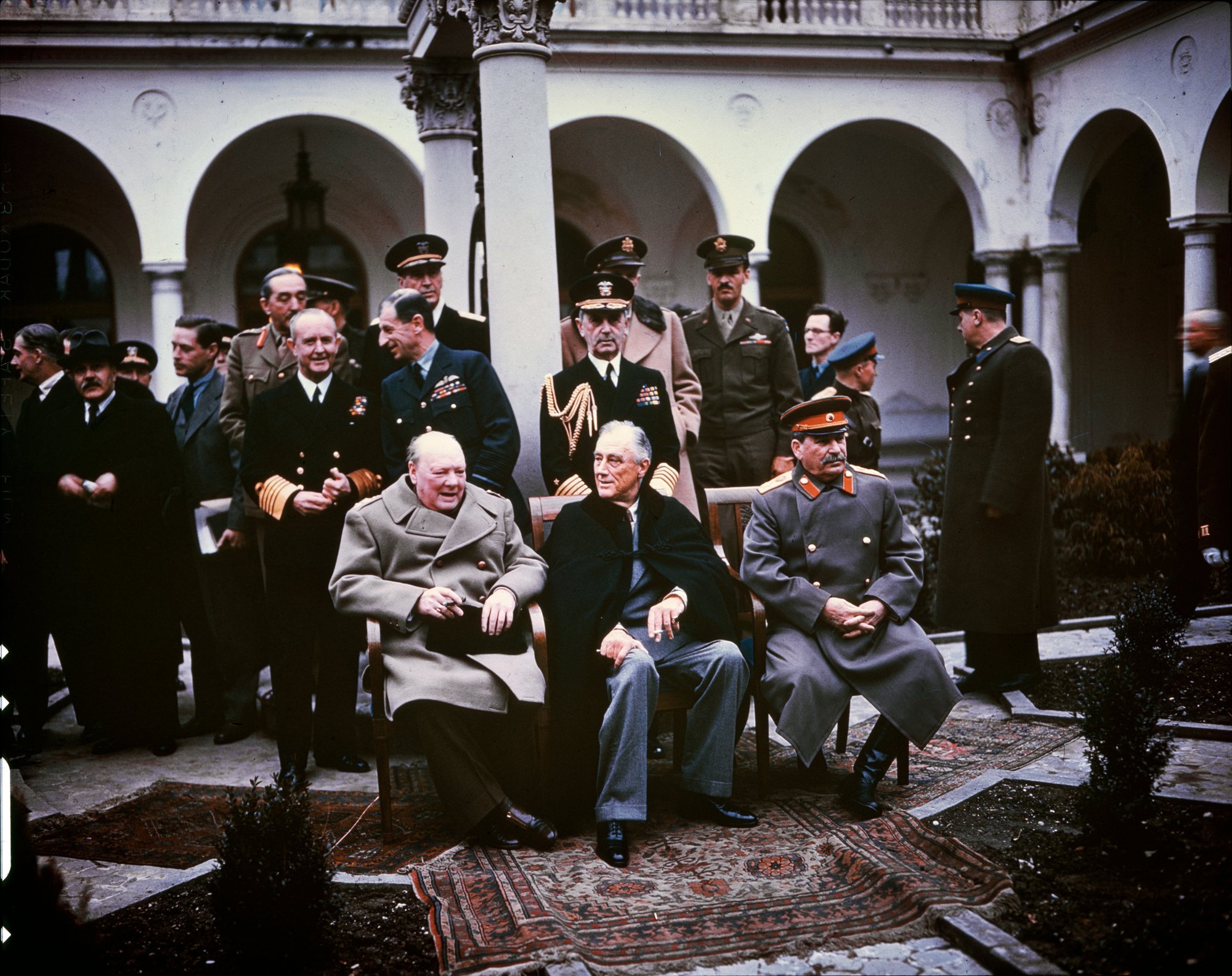Photographie en couleur prise à Yalta en février 1945. Au premier plan, Winston Churchill (à gauche), Franklin Roosevelt assis (au centre) et Joseph Staline (à droite) posent côte à côte. Derrière eux, des conseillers militaires.